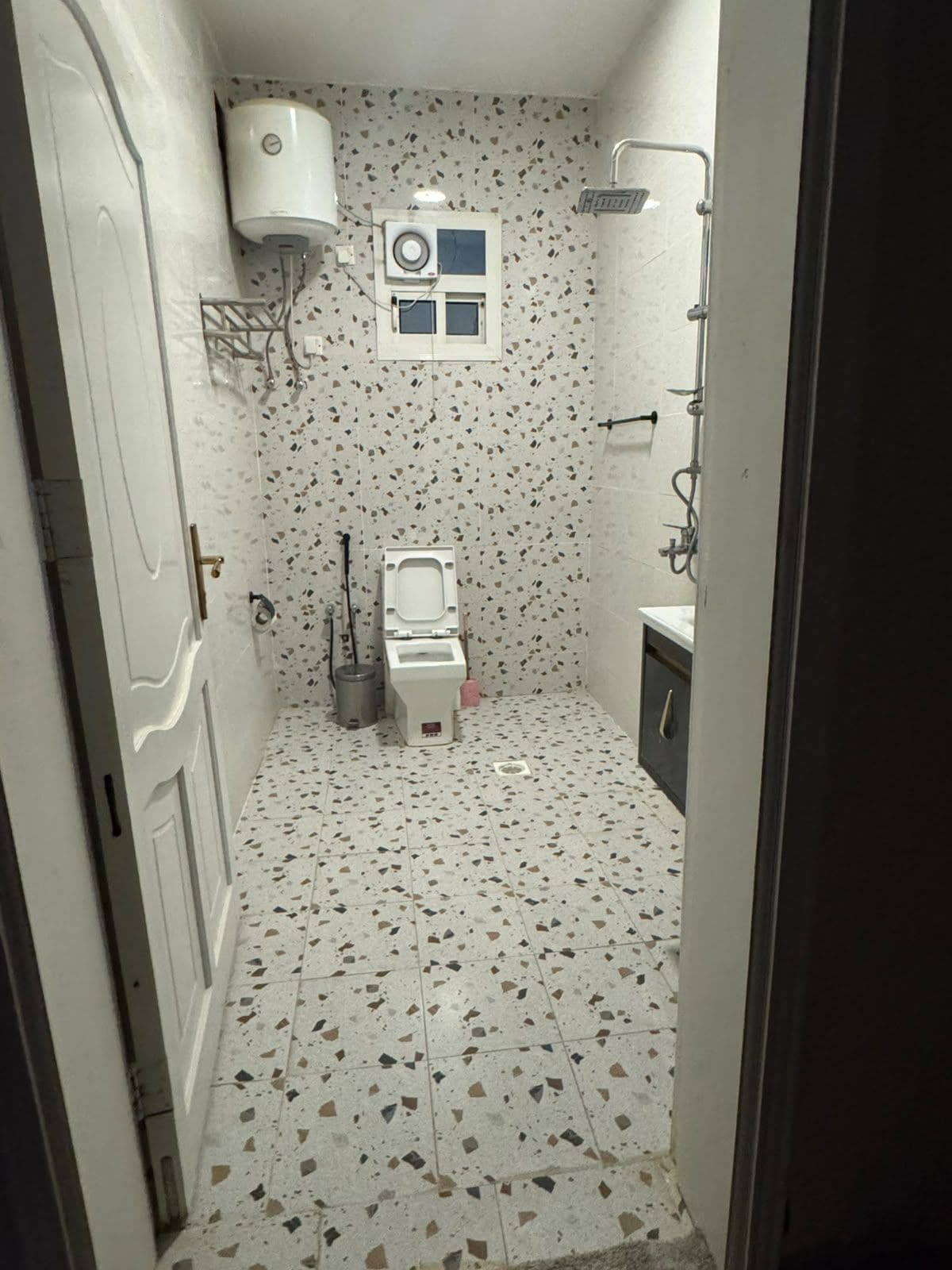 Un baño pequeño con inodoro, lavabo y ducha. Hay una ventana sobre el inodoro y un calentador de agua montado en la pared.
