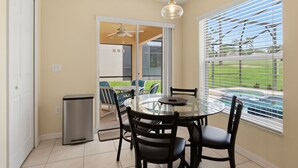 Una mesa de comedor con cubierta de vidrio, cuatro sillas, un ventilador de techo y vistas a una piscina a través de amplios ventanales.