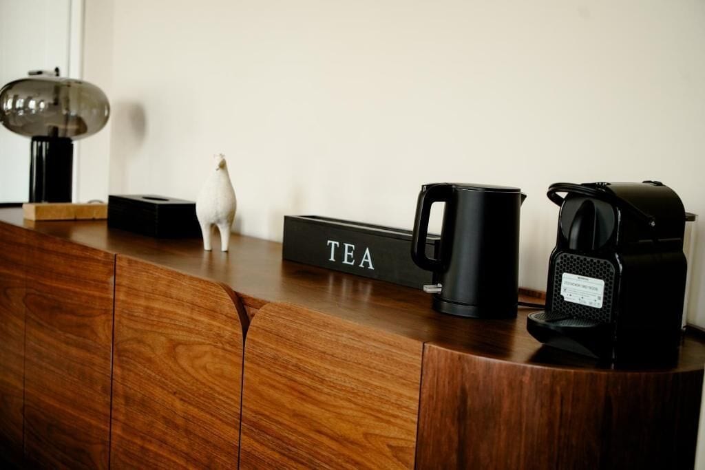 A wooden counter with a black box labeled 'TEA,' a black coffee maker, and a small white figurine.
