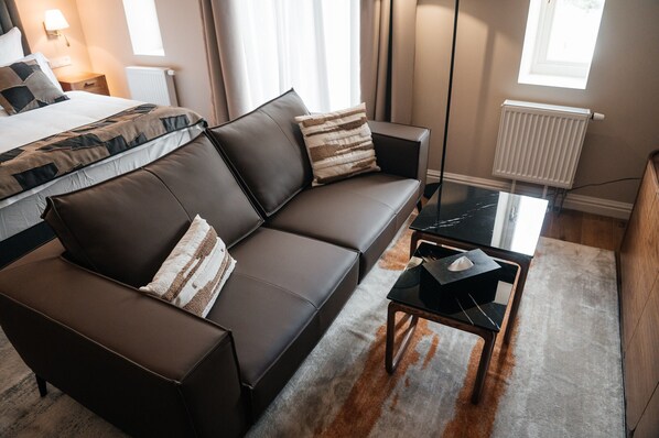 Una habitación de hotel moderna con un sofá de cuero marrón oscuro, una mesa de centro de vidrio y una cama con ropa de cama estampada.