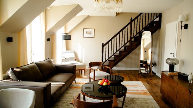A spacious living room with a brown leather sofa, a round coffee table with fruit, a wooden staircase, and a chandelier.