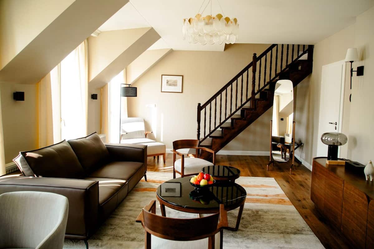 A spacious living room with a brown leather sofa, a round coffee table with fruit, a wooden staircase, and a chandelier.
