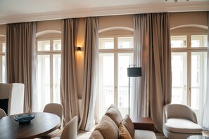 Quarto de hotel com sofá, cadeiras, mesa de jantar e janelas amplas com cortinas.