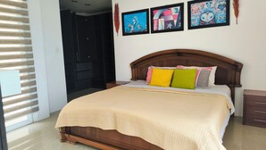 A bedroom with a wooden bed, a beige quilt, and colorful pillows. There are three framed artworks on the wall.