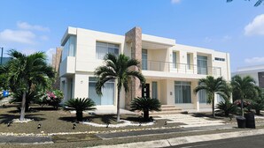 A modern two-story building with a balcony, surrounded by palm trees and a gravel driveway.