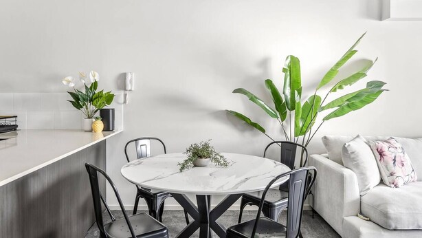 En moderne stue med et rundt spisebord, sorte stole, en hvid sofa med blomstermønstrede puder og store grønne planter.