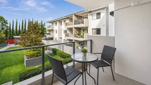 En balkon med et glasbord og to stole, med udsigt over en boligbygning med flere balkoner og en velplejet have.