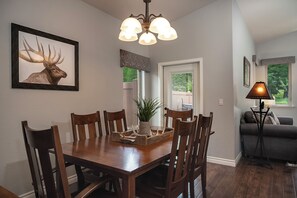 A dining area with a wooden table, chairs, a chandelier, a framed deer artwork, a potted plant, and a window with a view.