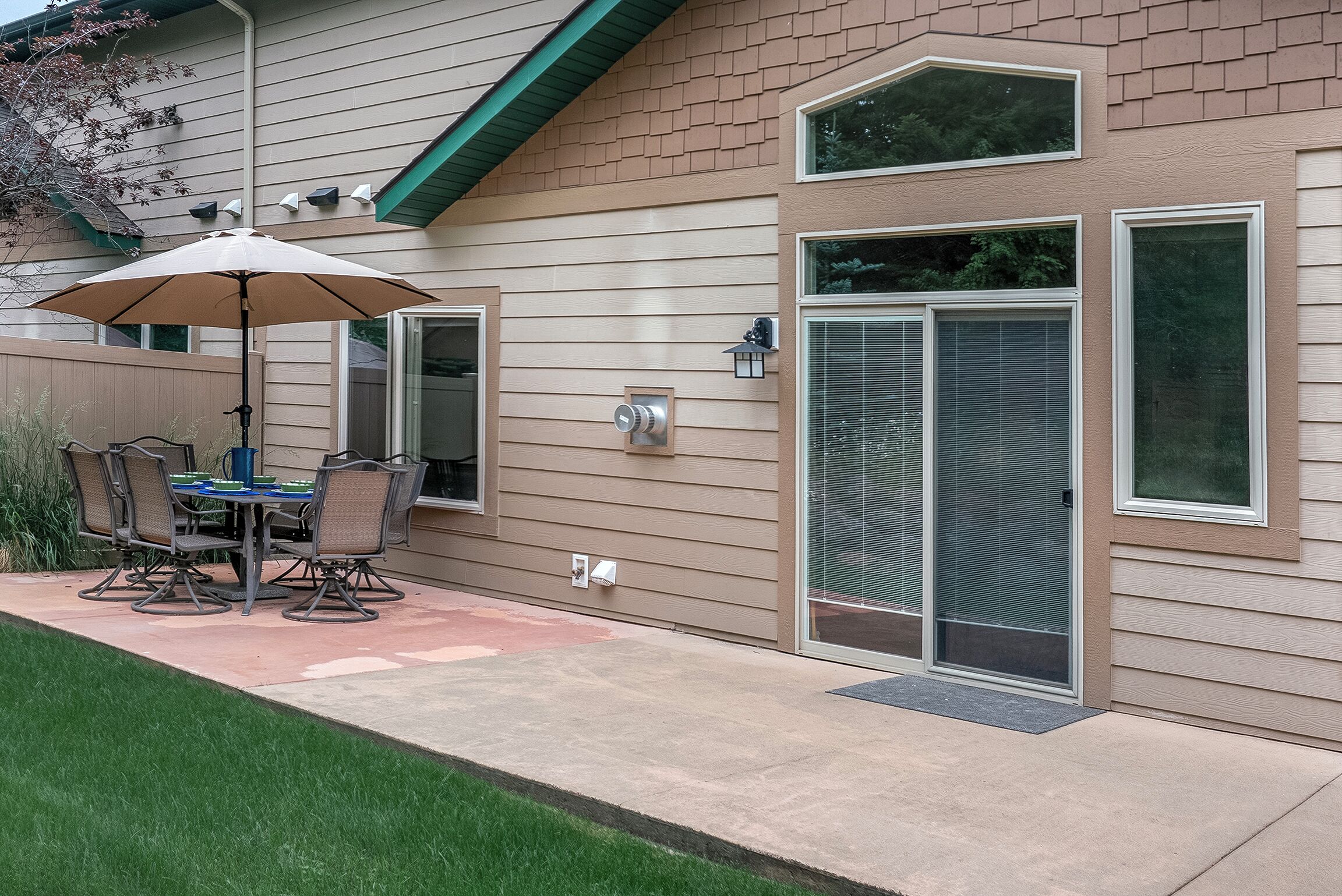 A patio with a table and chairs, a covered umbrella, and a house with a garage door and a window.