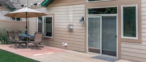 A patio with a table and chairs, a covered umbrella, and a house with a garage door and a window.