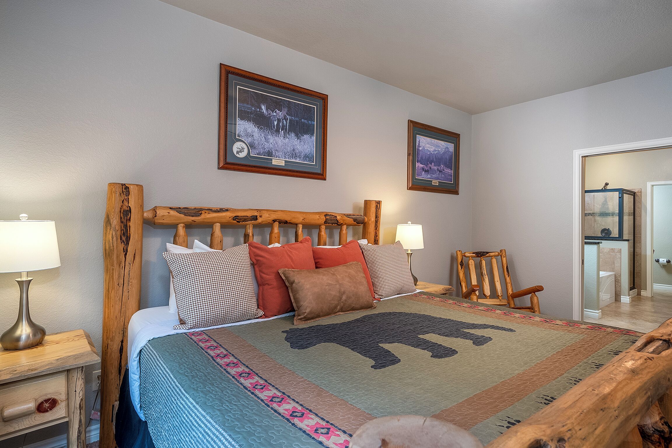 Una cama con cabecera de madera, dos cuadros enmarcados en la pared, una silla de madera y una mesita de noche con una lámpara.