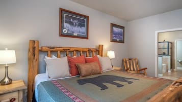 Una cama con cabecera de madera, dos cuadros enmarcados en la pared, una silla de madera y una mesita de noche con una lámpara.