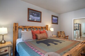 Una cama con cabecera de madera, dos cuadros enmarcados en la pared, una silla de madera y una mesita de noche con una lámpara.
