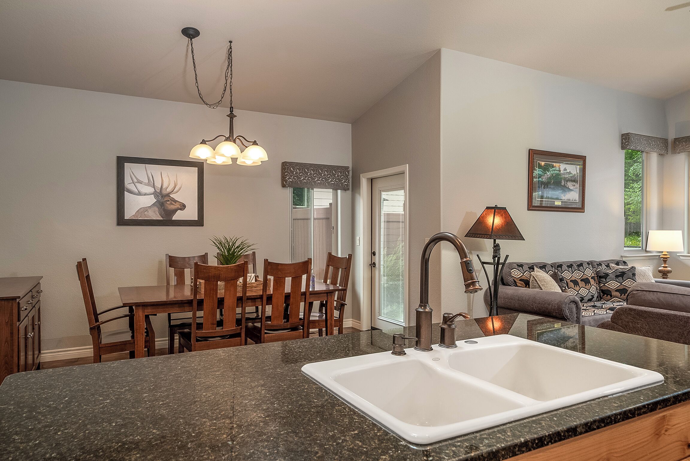 A kitchen with granite countertops, a double sink, wooden dining table, chairs, and a chandelier.