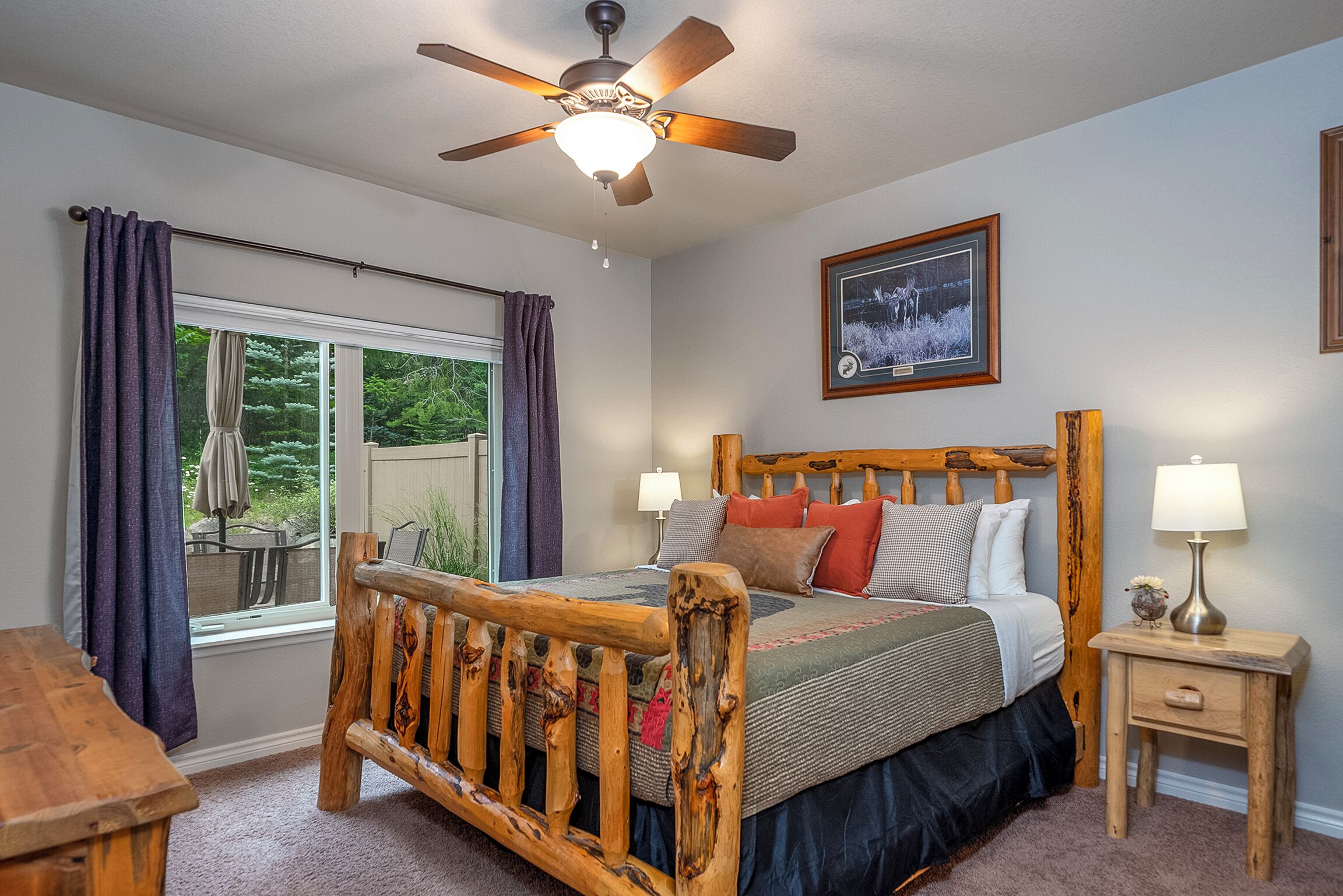 A bedroom with a wooden bed frame, a ceiling fan, a window with curtains, a bedside table with a lamp, and a framed picture on the wall.