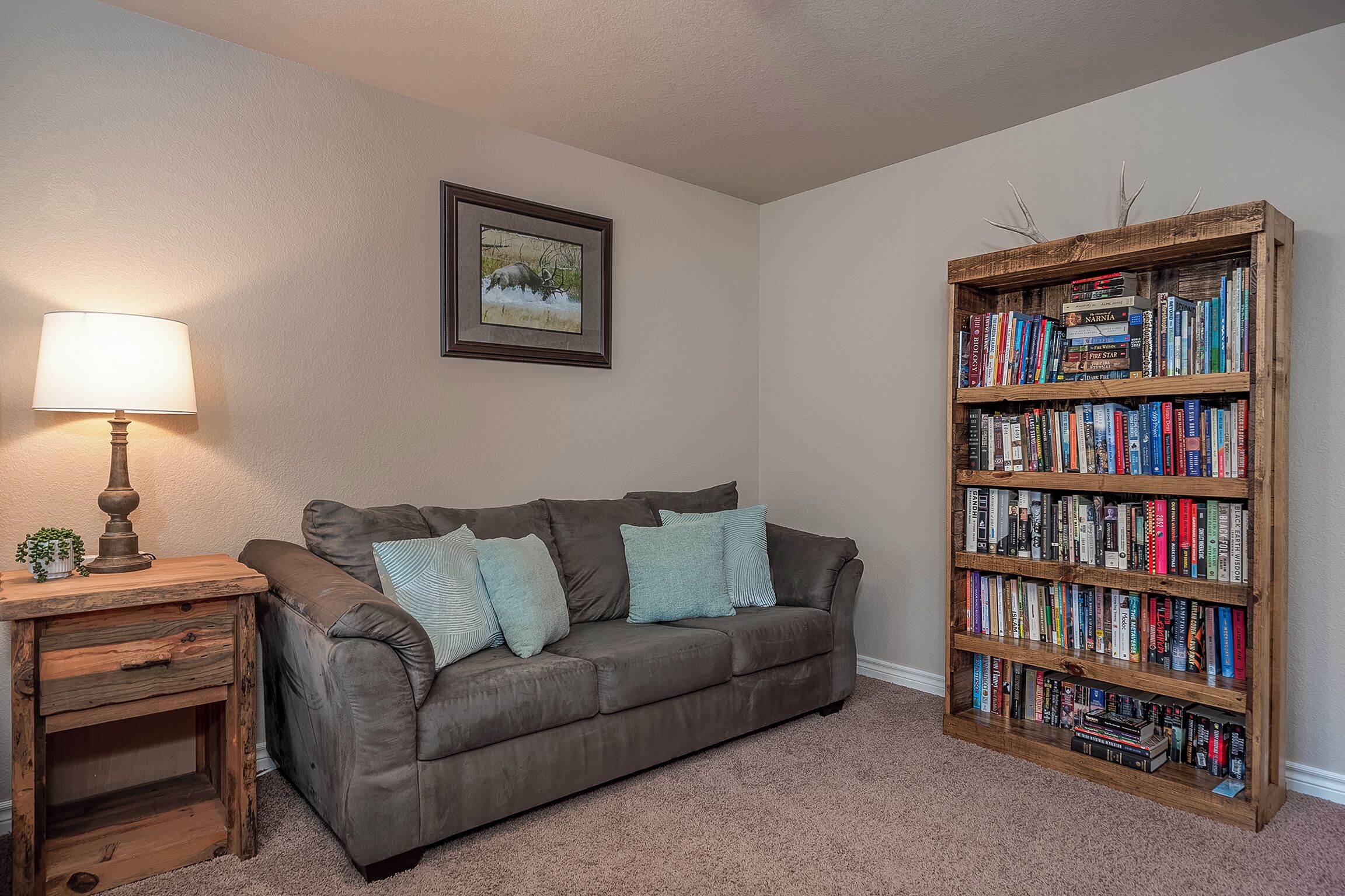 Un salon avec un canapé gris, une table d’appoint en bois surmontée d’une lampe, un tableau encadré accroché au mur et une bibliothèque remplie de livres.