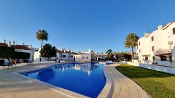 A swimming pool with a tiled deck, surrounded by white buildings with red roofs, palm trees, and a clear blue sky.