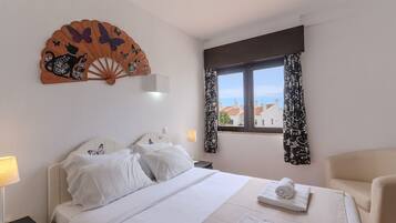A hotel room with a bed, a chair, a window with curtains, a wall fan, and a view of buildings.