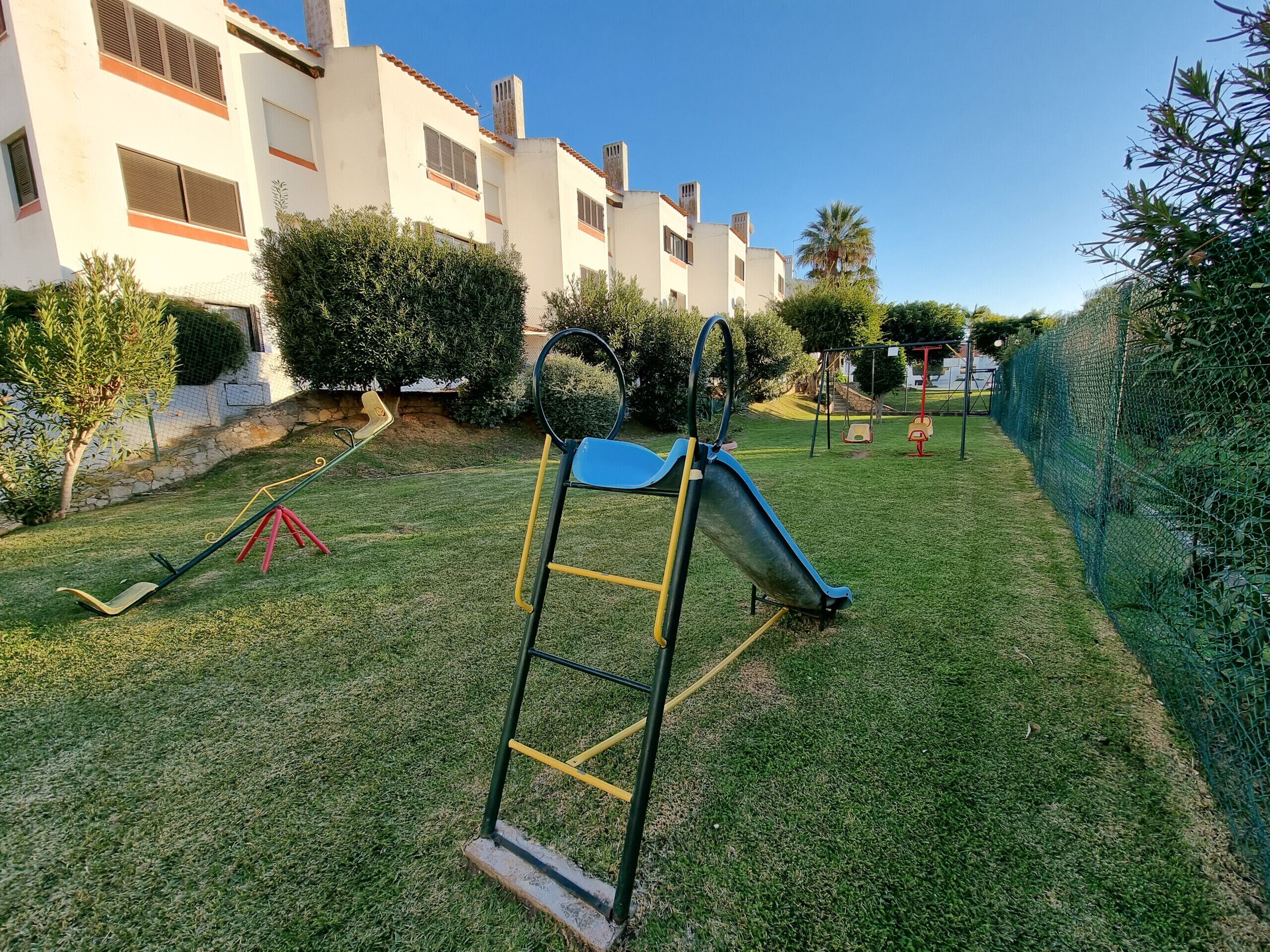 Área residencial com parquinho, incluindo escorregador e balanços, cercada por vegetação e uma grade.
