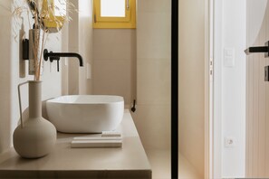 Ein Badezimmer mit einem weißen Waschbecken, einer Vase mit Trockenpflanzen und einem gelben Fenster.
