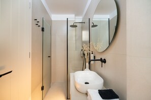 A modern bathroom with a glass shower enclosure, a white sink, and a round mirror. - Deluxe Suite with Shared Pool and Seaview (Kimolos)