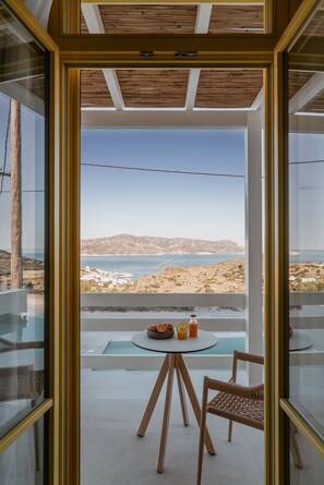A modern room with a glass wall, a round table with a chair, and a view of the sea and hills. - Deluxe Suite with Shared Pool and Seaview (Kimolos)