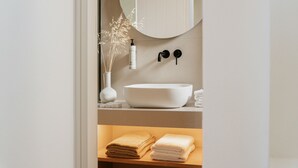Ein Badezimmer mit einem weißen Waschbecken, einem Spiegel, einer Vase mit Trockenpflanzen und Handtüchern auf einem Regal.