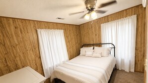 Un dormitorio con cama, ventilador de techo, paredes de madera y cortinas blancas.