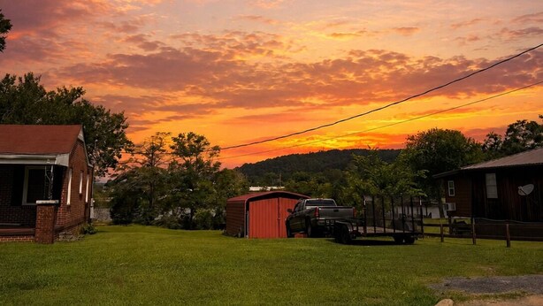 A rural landscape with a house, a red shed, and a truck parked on a grassy area.