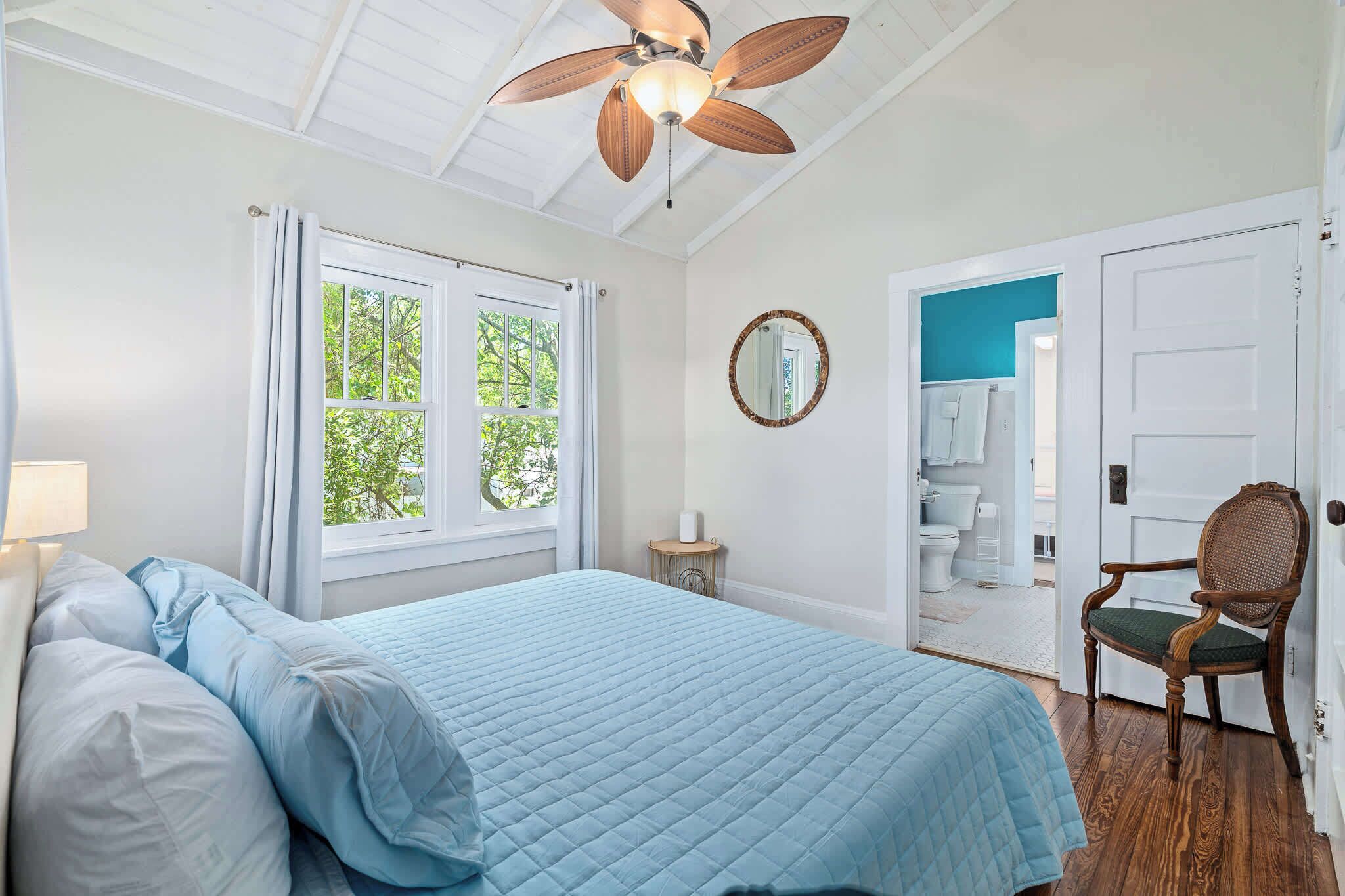 Un dormitorio con cama, ventilador de techo, ventana, silla y puerta que da a un baño.