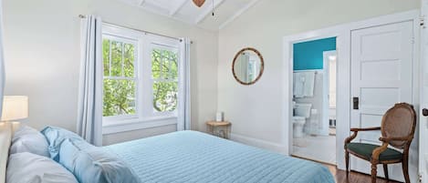 Un dormitorio con cama, ventilador de techo, ventana, silla y puerta que da a un baño.