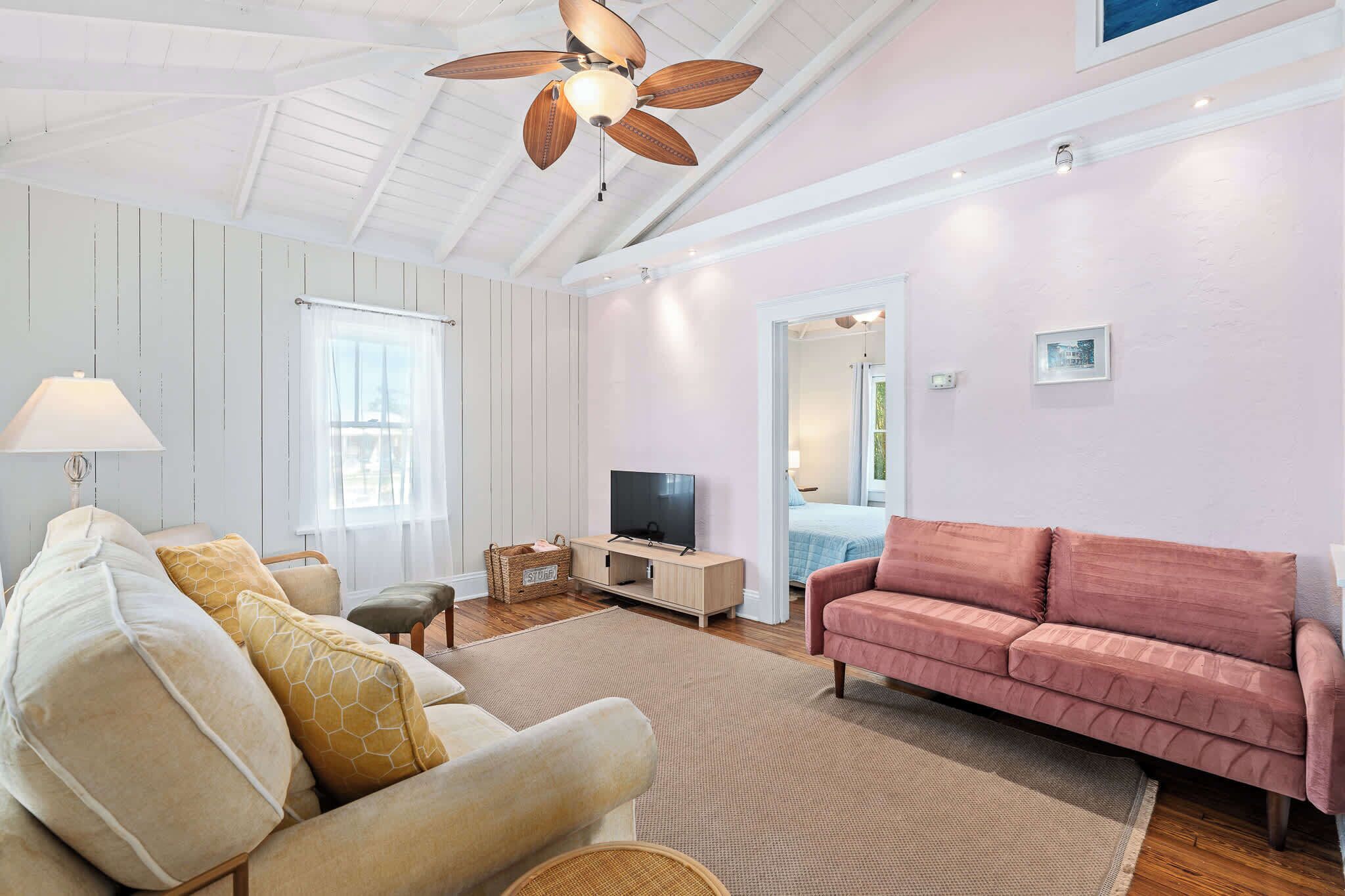Una sala de estar con ventilador de techo, televisor, sofá rosa y sofá blanco.