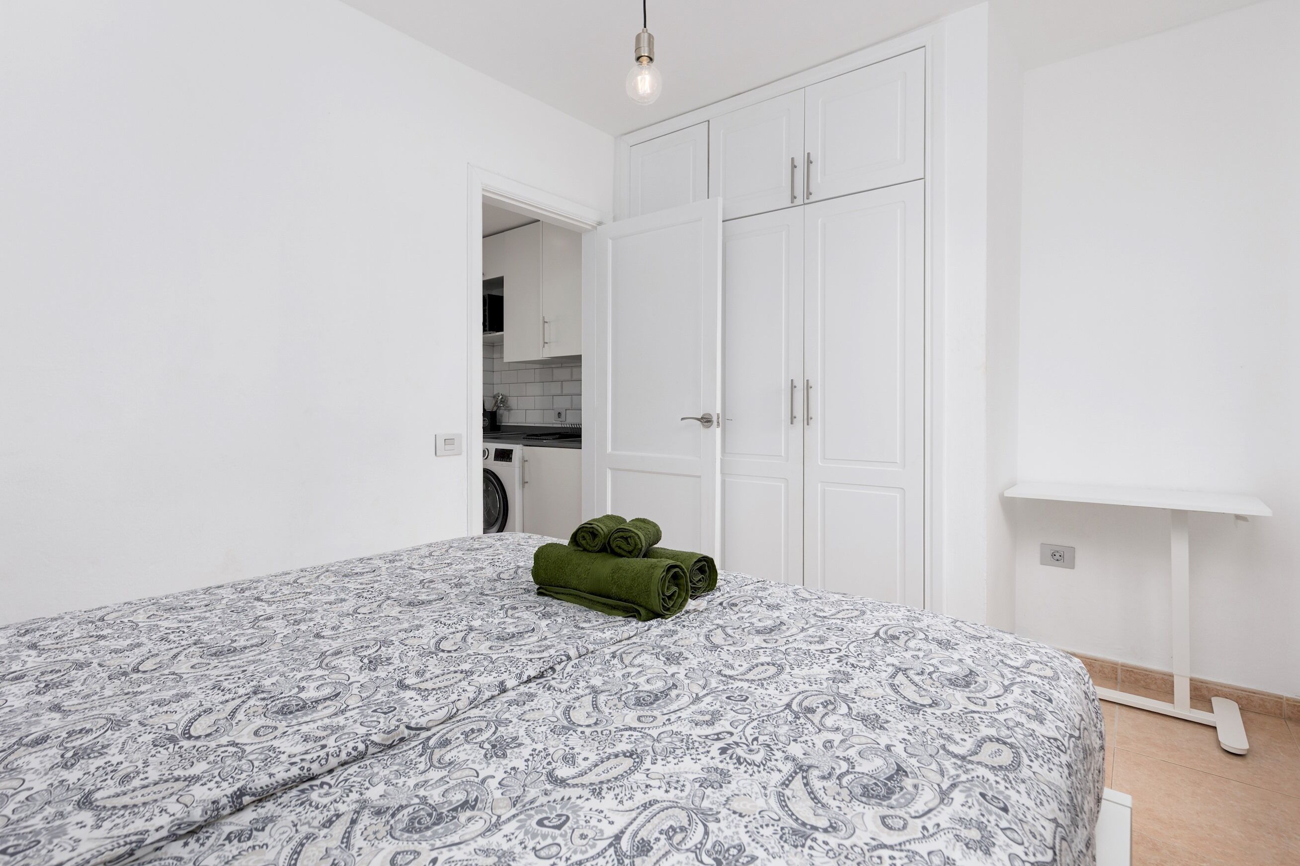 Une chambre avec une couvre-lit à motifs, une serviette verte, des armoires blanches et un coin cuisine visible.