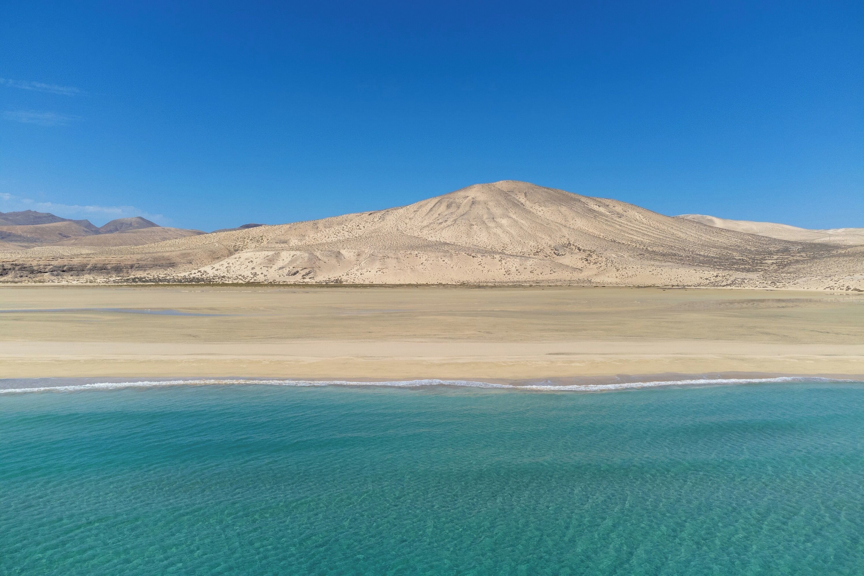 A desert landscape with a mountain in the background and a clear blue sea in the foreground.