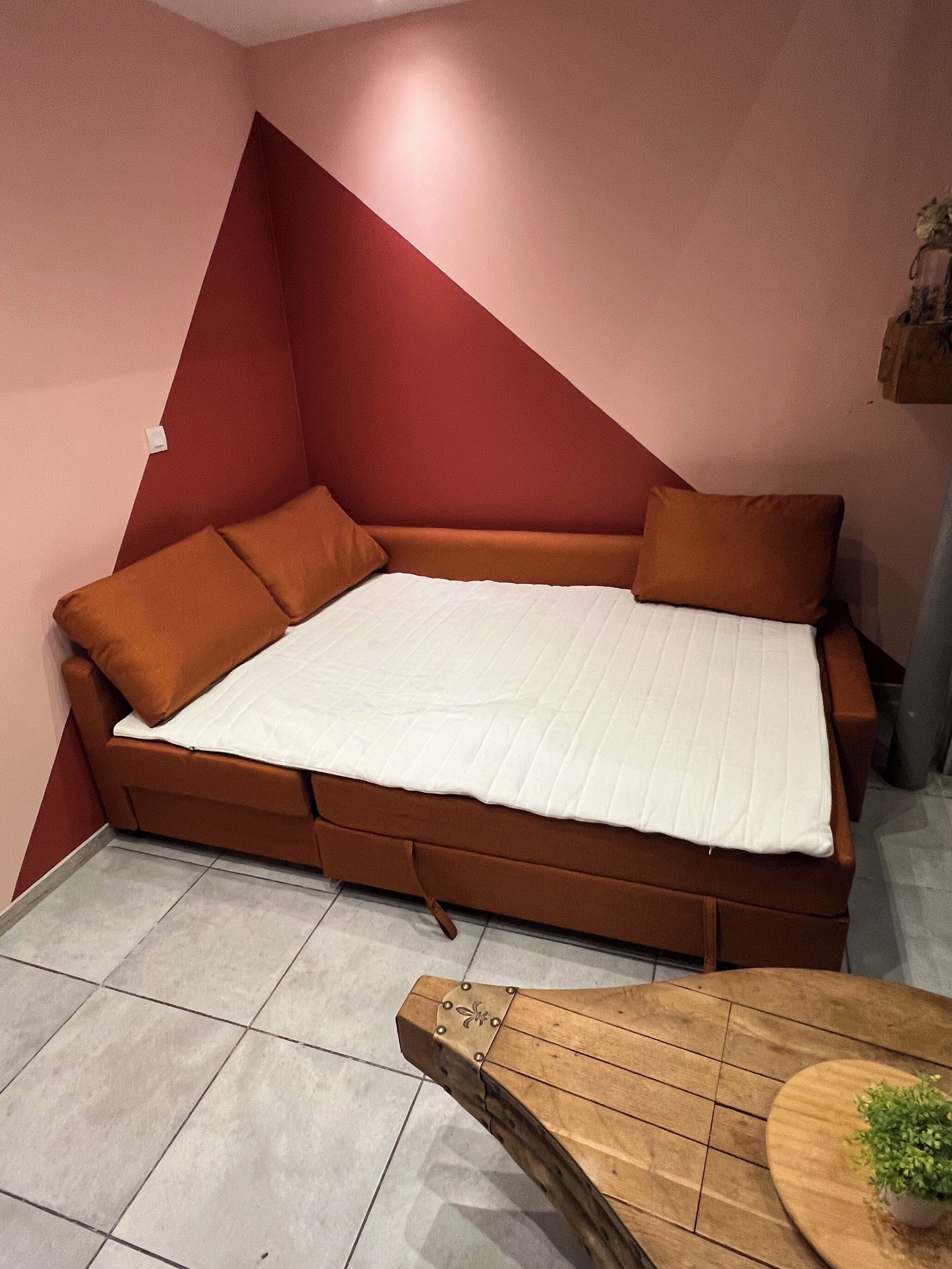 Een hoekbed met een witte matras, een bruin hoofdeinde en bijpassende kussens, in een kamer met een betegelde vloer en een houten tafel met een plant.