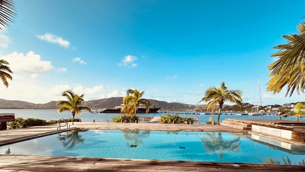 Zona de piscina con agua cristalina, muelle de madera, palmeras y una vista distante de montañas y agua.
