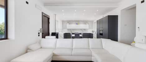 A modern living room with a white sectional sofa, a dining area with a table and chairs, and a kitchen area with cabinets and appliances.
