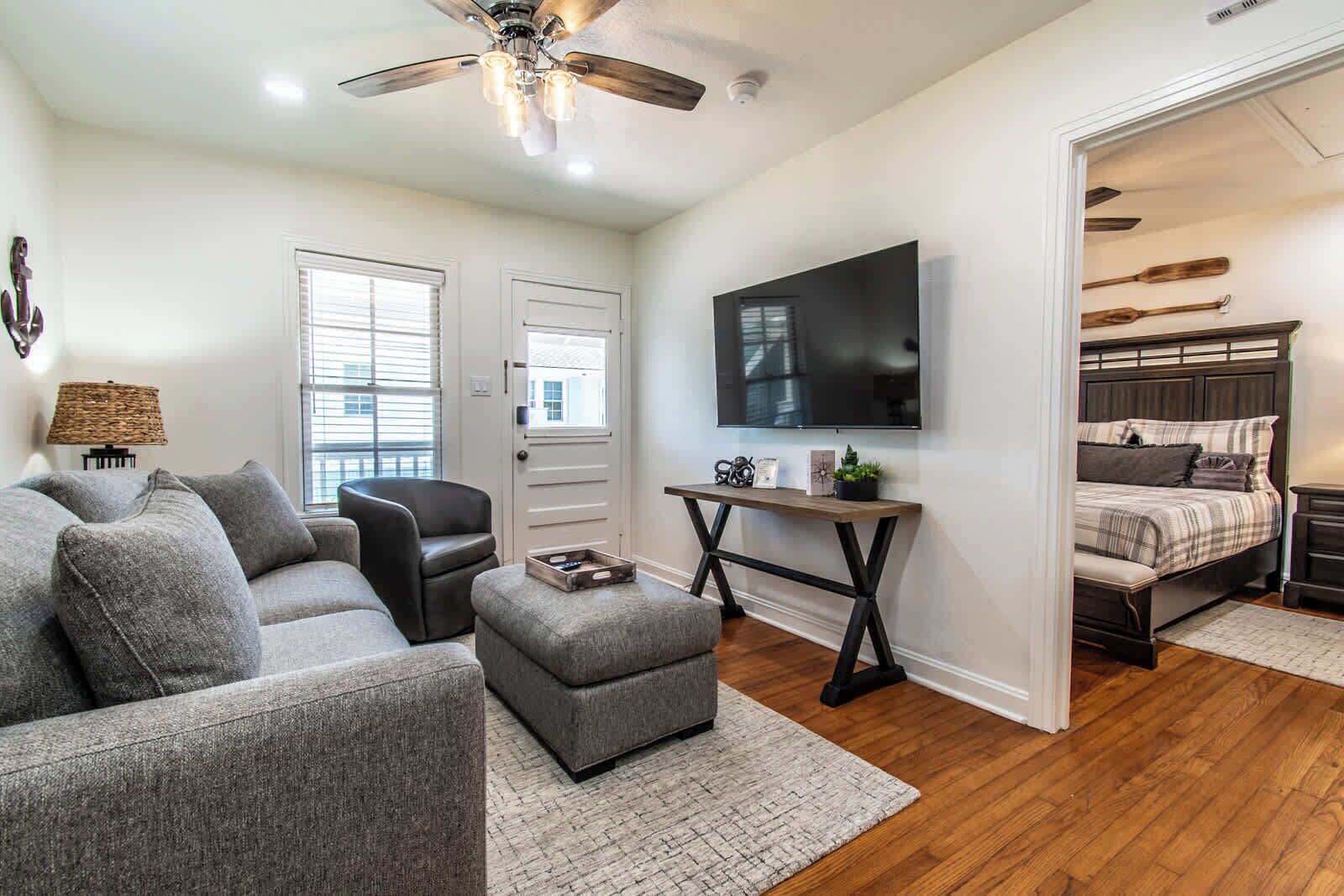 A modern living room with a grey sofa, a small armchair, a coffee table, a TV mounted on the wall, and a wooden floor.