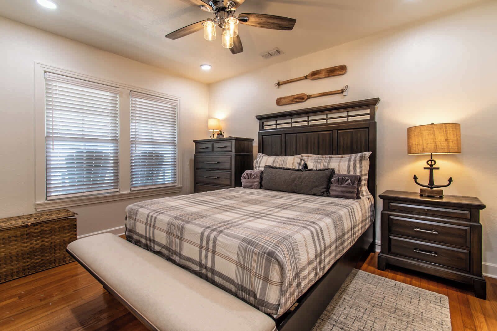 A bedroom with a bed, bedside tables, a bench, a ceiling fan, and a window with blinds.