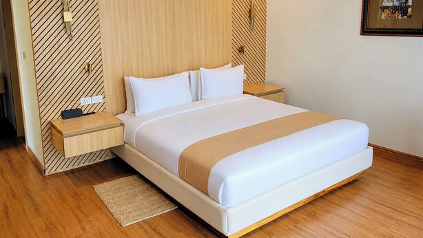 Una cama bien hecha con cabecera de madera, una mesita de noche con lámpara y una alfombra pequeña sobre el suelo de madera.