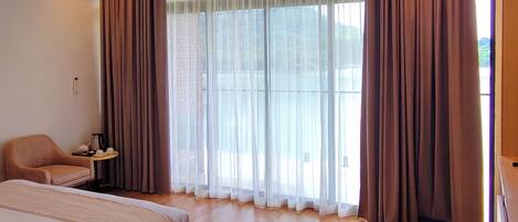 A hotel room with a bed, a chair, a small table with a lamp, and a large window with curtains.