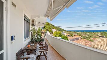 A balcony with a table and chairs, overlooking a coastal town with houses and a view of the sea.