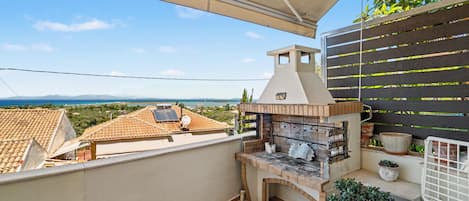 A balcony with a barbecue grill, potted plants, and a view of the sea and mountains.