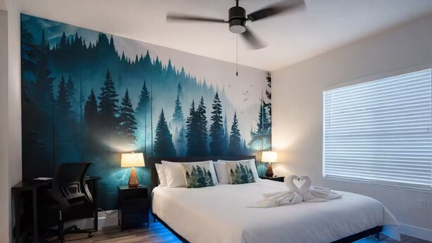 Ein Schlafzimmer mit einem Bett, zwei Nachttischlampen, einem Schreibtisch mit Stuhl, einem Deckenventilator und einem großen Wandgemälde von einem Wald.