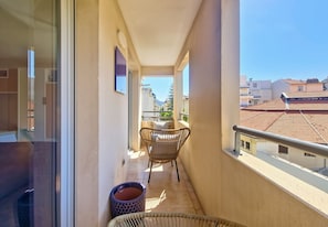 Un balcón con una silla y una mesa pequeña, una ventana con vistas a edificios, y un cubo de basura.