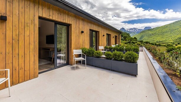 Un bâtiment moderne aux murs extérieurs en bois, doté d’un balcon avec des chaises et offrant une vue sur une vallée verdoyante et des montagnes.