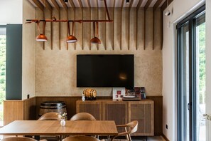 A modern interior with a wooden dining table, chairs, a flat-screen TV mounted on a textured wall, and a wooden cabinet with a barrel and decorative items. - Odat n`Bogë | Room 2 - Bjeshka | Breakfast Included by PikHost (Bogë)
