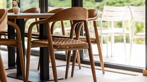 A dining area with wooden chairs and a table, large windows with a view of greenery, and a white chair outside. - Odat n`Bogë | Room 2 - Bjeshka | Breakfast Included by PikHost (Bogë)