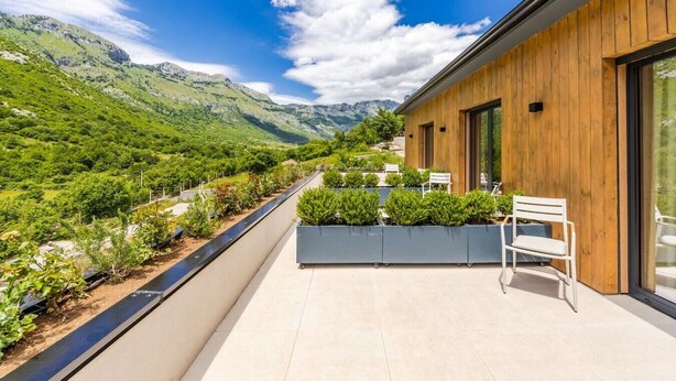 Een modern gebouw met een houten gevel, een balkon met zithoek, en uitzicht op een groene vallei en bergen.