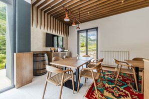 A modern dining area with a wooden table, chairs, and a large window offering a view of the outdoors. - Odat n`Bogë | Room 3 - Mali | Breakfast Included by PikHost (Bogë)